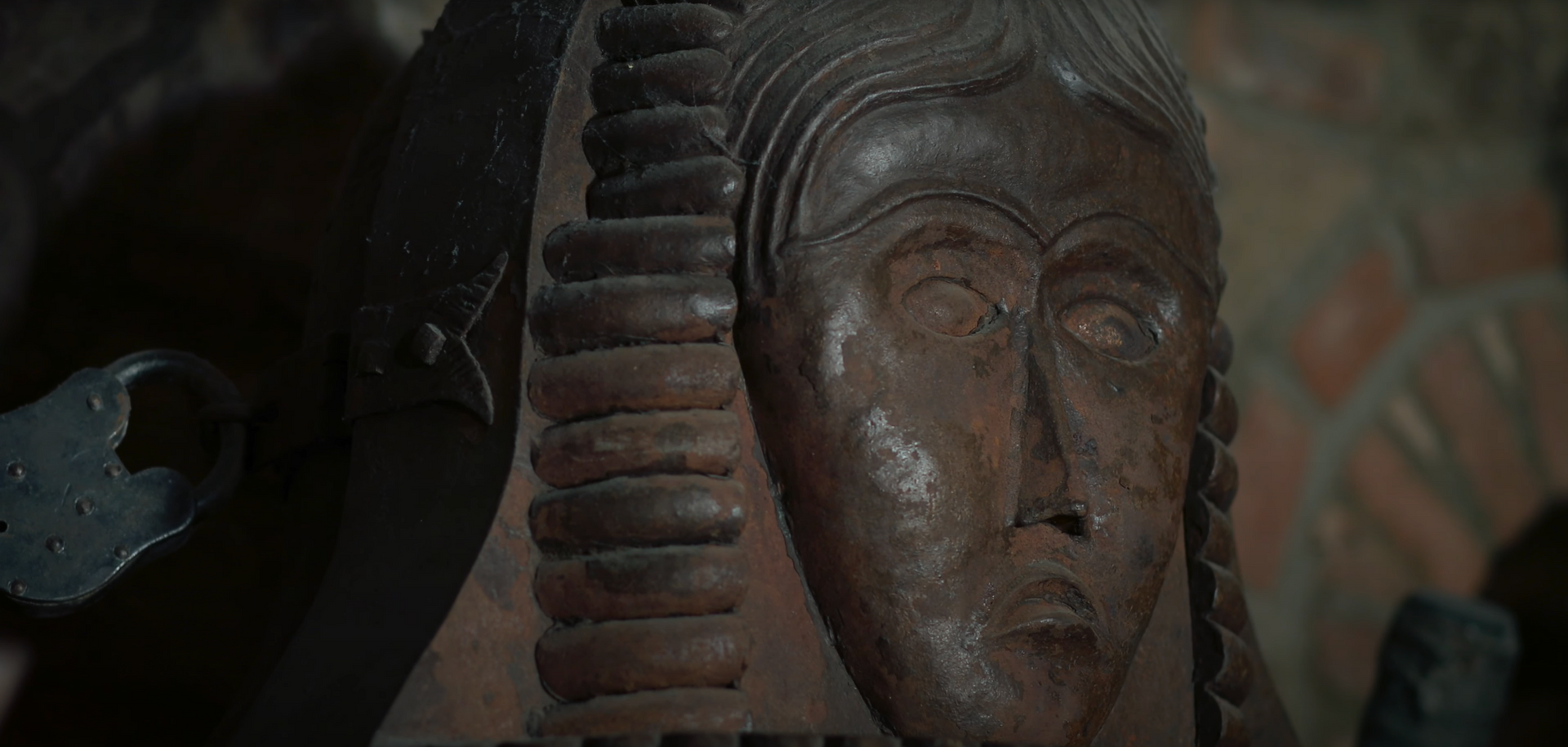 Carved wooden face with braided hair, set in a dimly lit, possibly ancient interior.