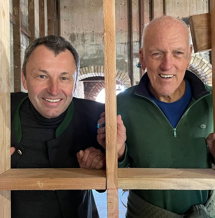 Two smiling men looking through a wooden frame in a partially constructed room.