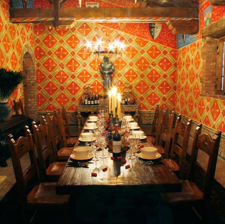 Dining room with long wooden table set for a feast; orange patterned walls, candles, knight statue, and wooden chairs.