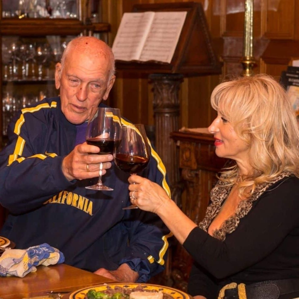 Man and woman toasting with red wine at a table. Wooden interior with music sheet and glass display.