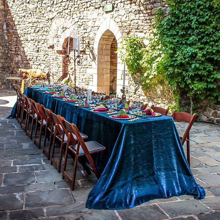 Long table set for a meal in a courtyard, covered in blue fabric, with chairs lined up.