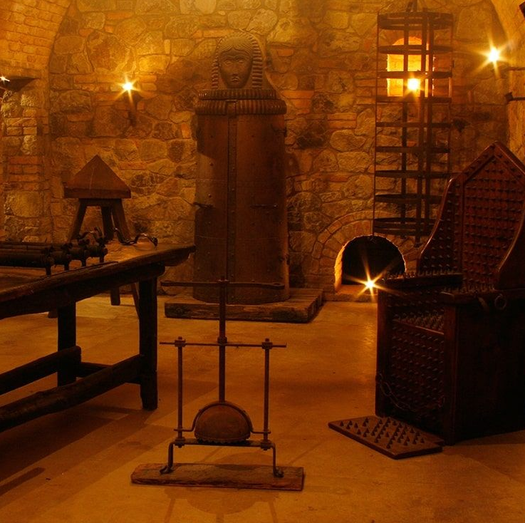 Dungeon interior with torture devices, including a rack, iron maiden, and spiked chair, lit by dim light.