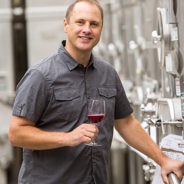 Man in gray shirt holds a glass of red wine, standing near stainless steel tanks.