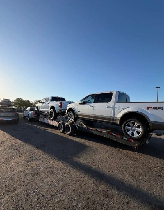 Two white pickup trucks loaded onto a flatbed car hauler trailer parked in an open lot under a clear blue sky.