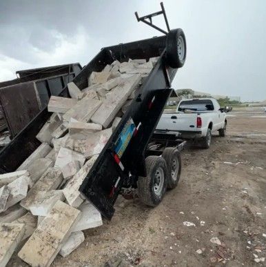 A truck bed being dumped, overflowing with concrete slabs, with a white pickup truck hitched in a dirt area.