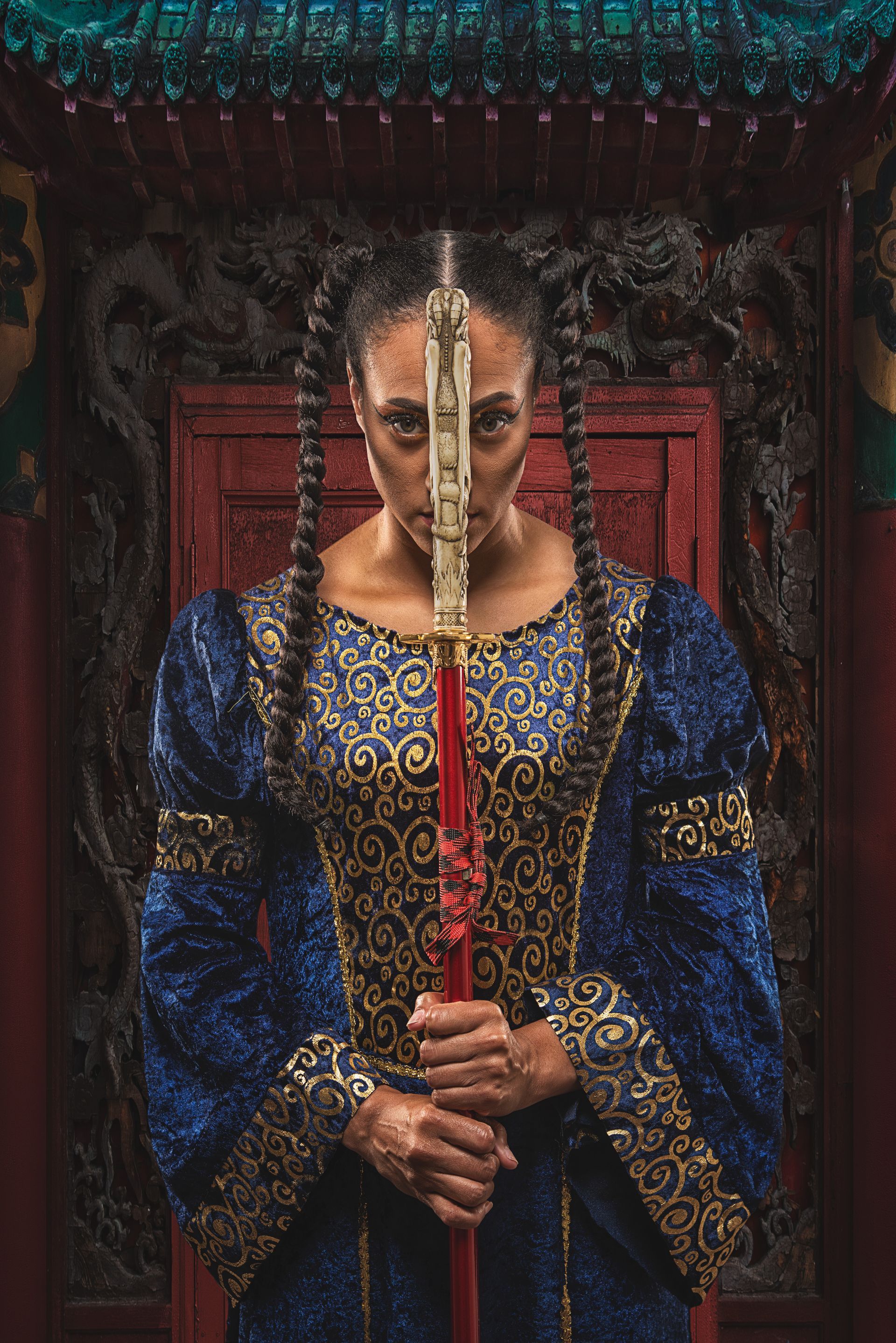 Woman holding a staff, facing camera. Blue and gold gown. Braided hair. Red and gold backdrop.