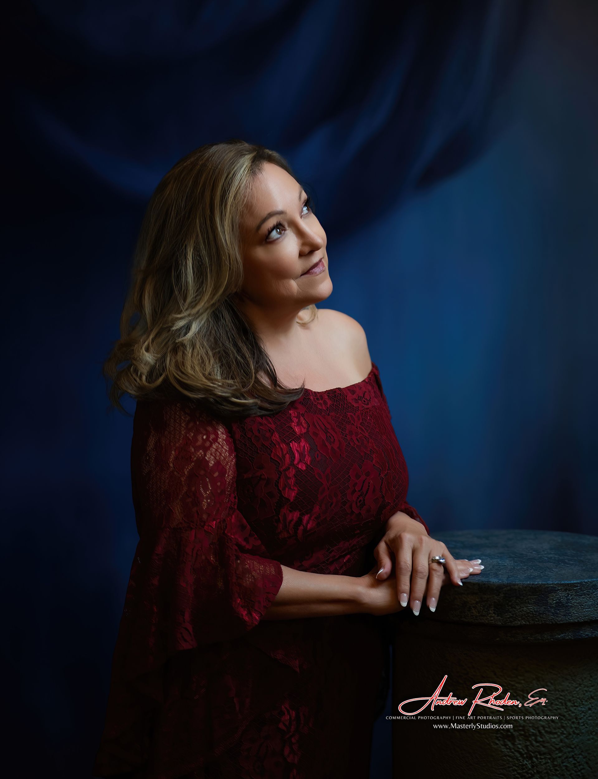 Woman in a red off-the-shoulder dress, looking up. Leaning on a stone surface, blue background.