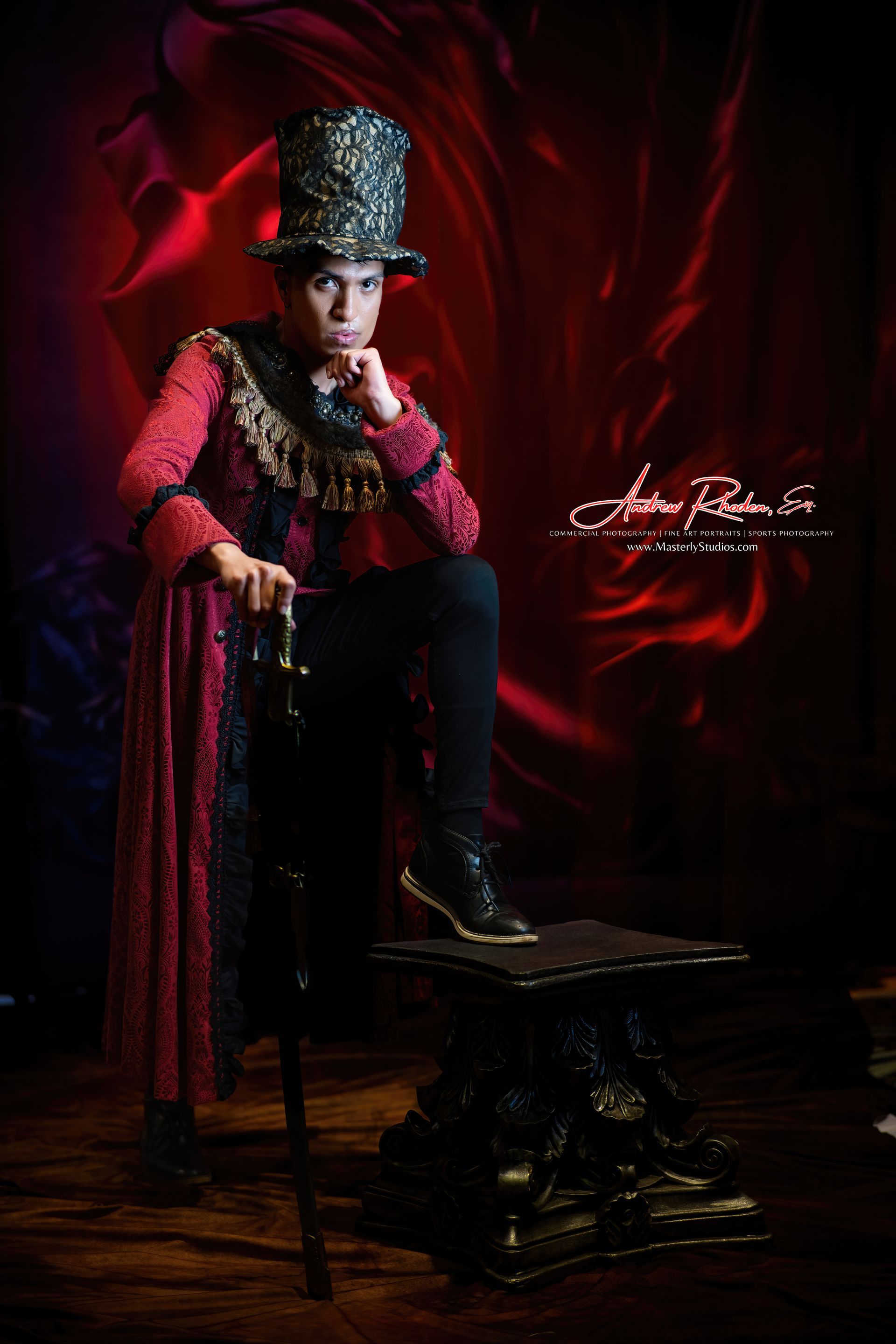 Person in elaborate red and black costume and top hat poses with cane, leg on a pedestal, against a red backdrop.