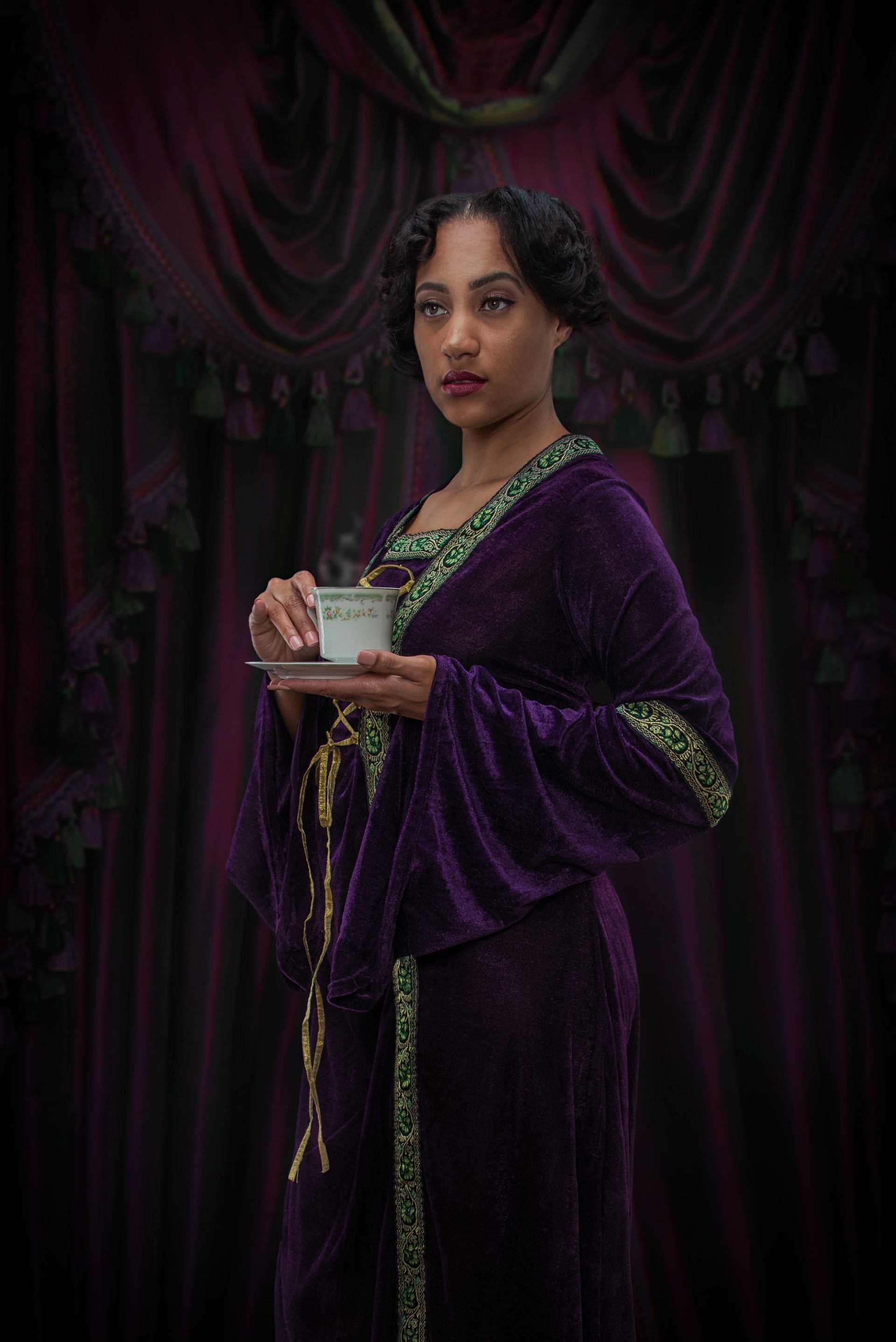 Woman in purple velvet gown holds teacup, stands before dark curtain.