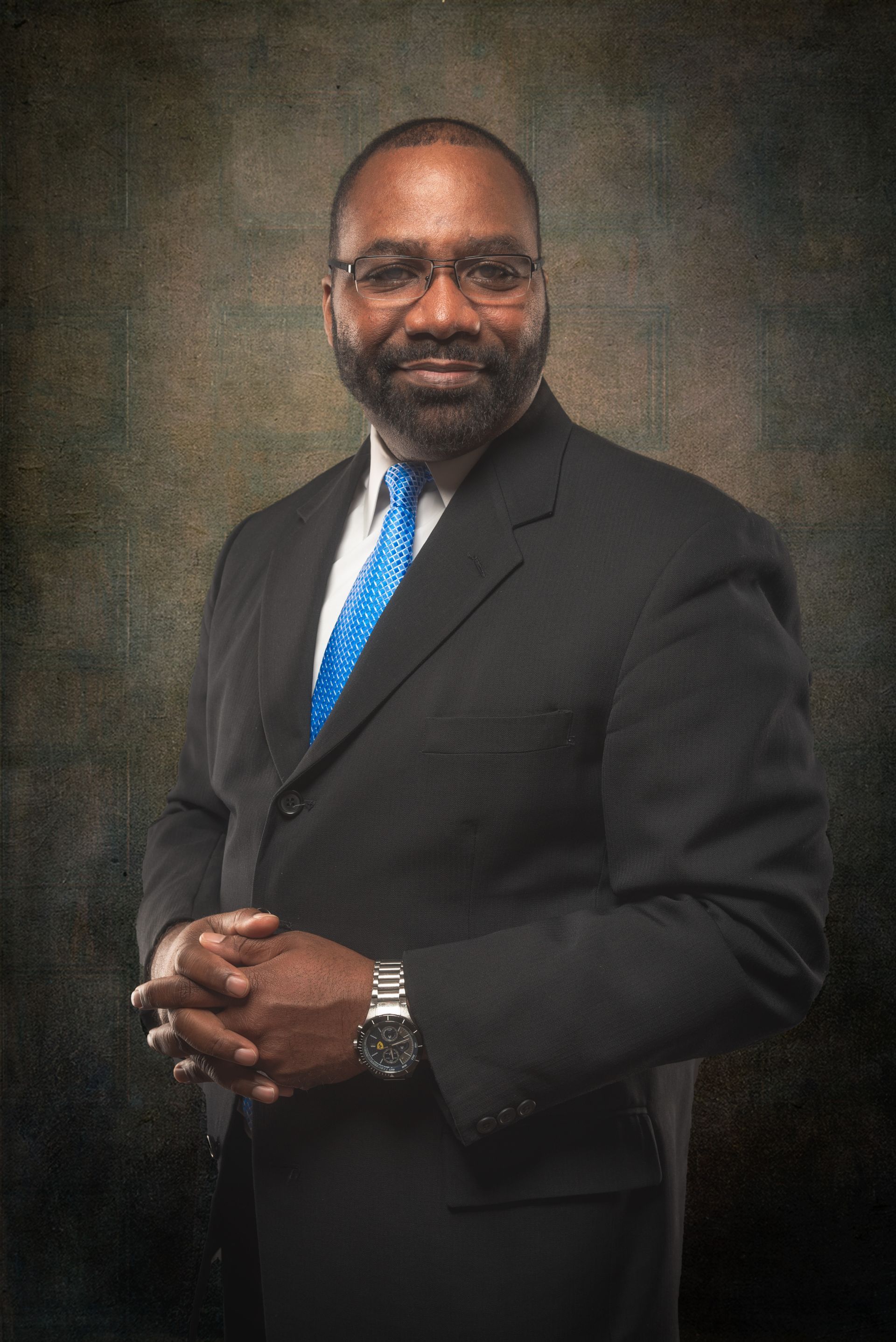 Man in a suit, hands clasped, smiling, against a textured brown and green background.
