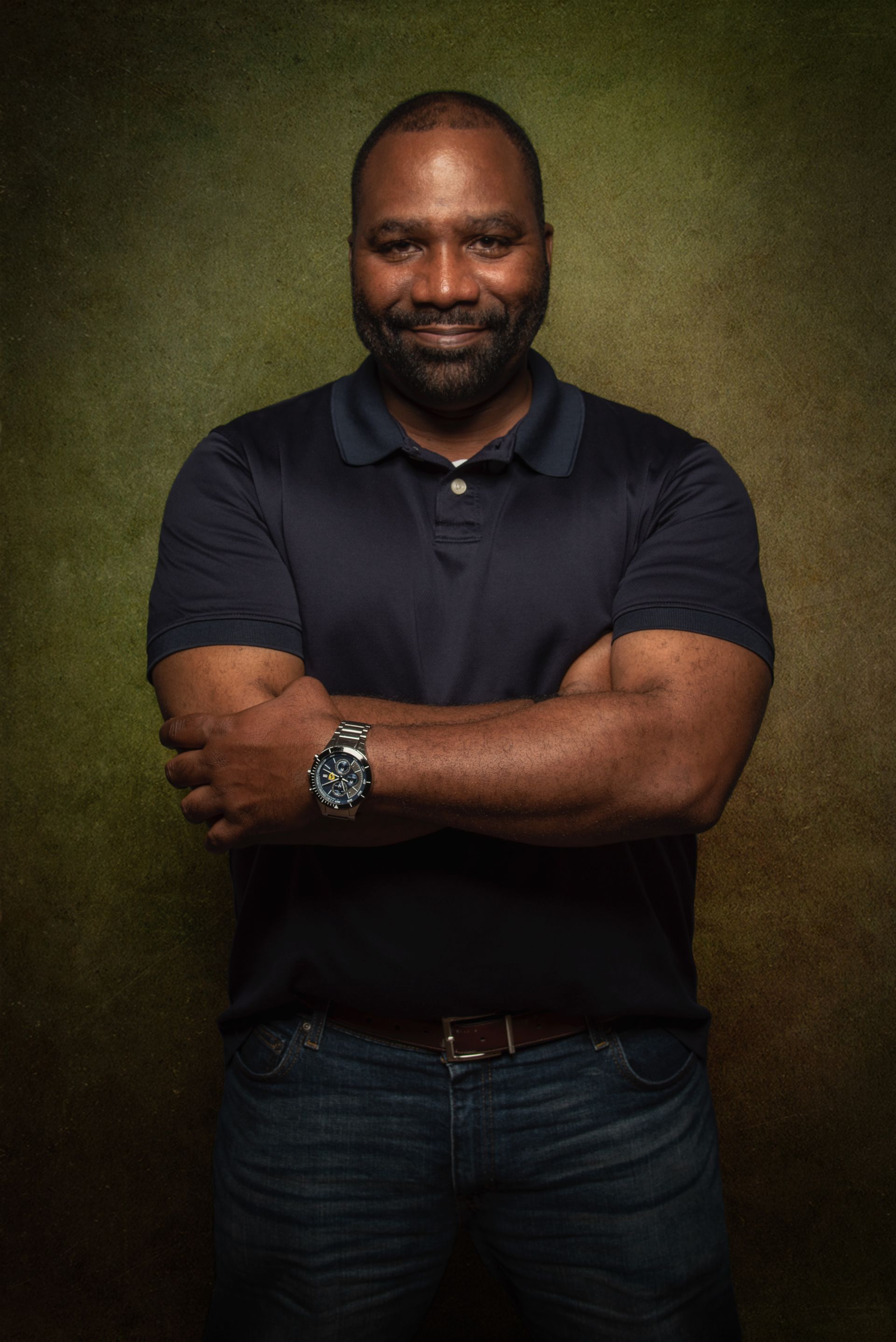 Man with arms crossed, wearing a navy polo shirt and jeans, standing against a green-toned backdrop.
