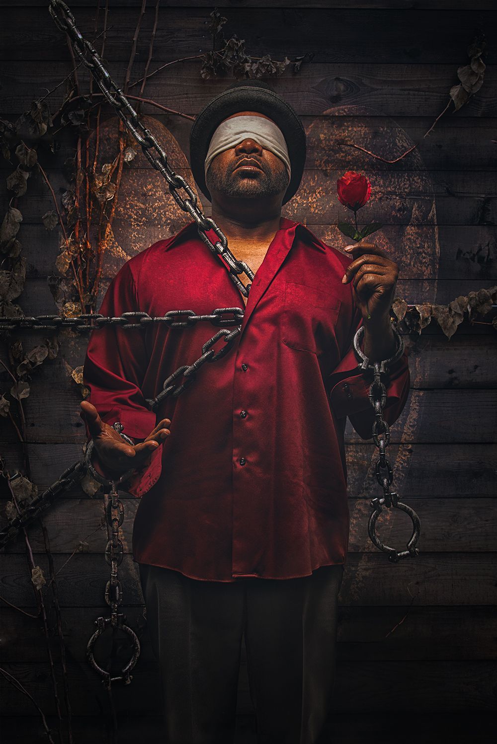 Man with blindfold, holding rose, chained. Red shirt, wooden background.