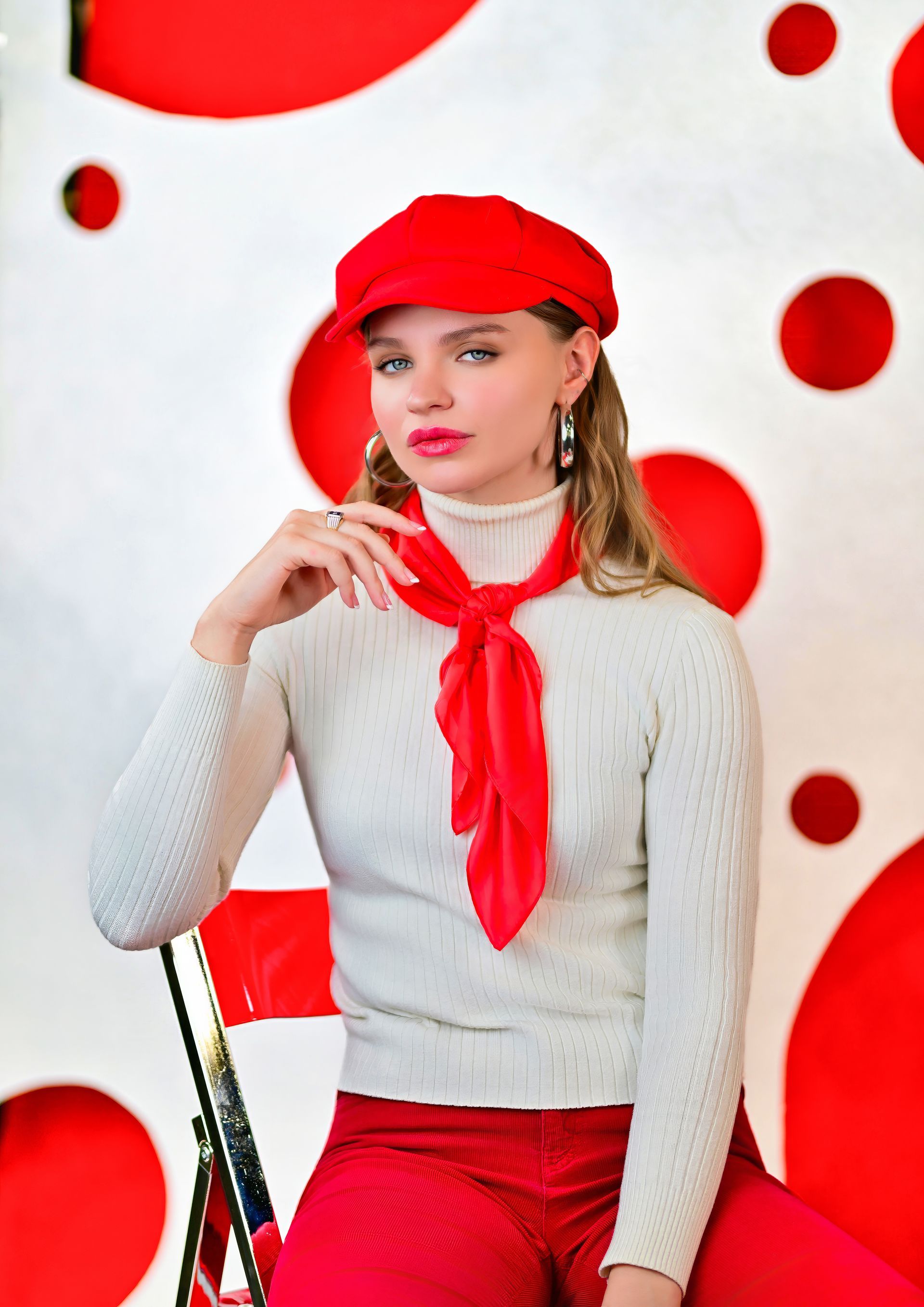 Woman in red hat, scarf, and pants sits, hand to face, against red-and-white backdrop.