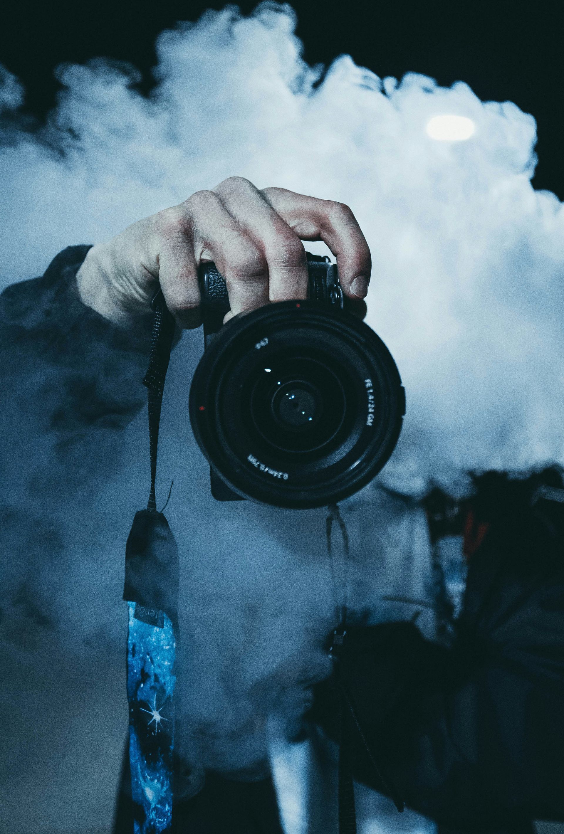 Hand holding a camera, lens pointed forward, surrounded by dense blue-gray smoke.