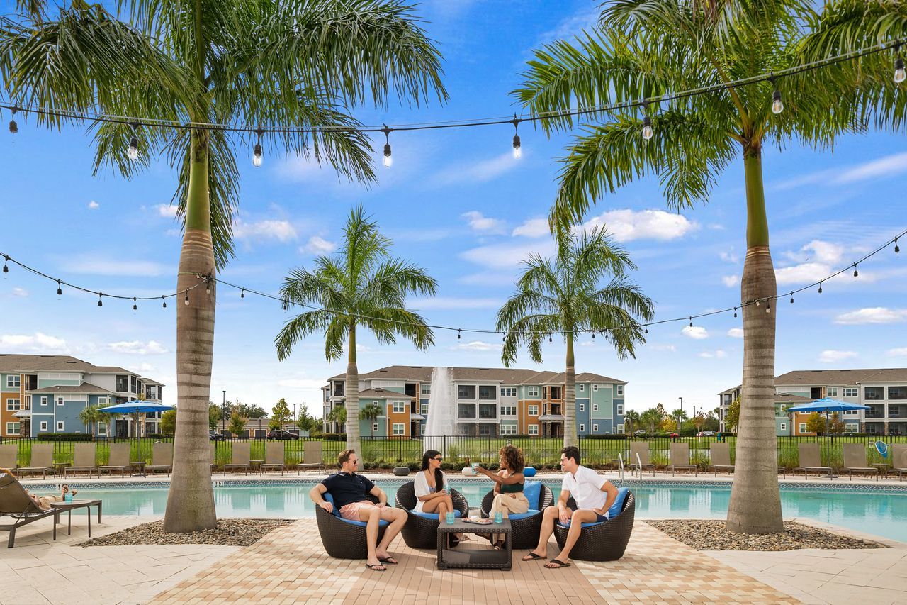 Group of friends lounging by pool under palm trees, blue sky.