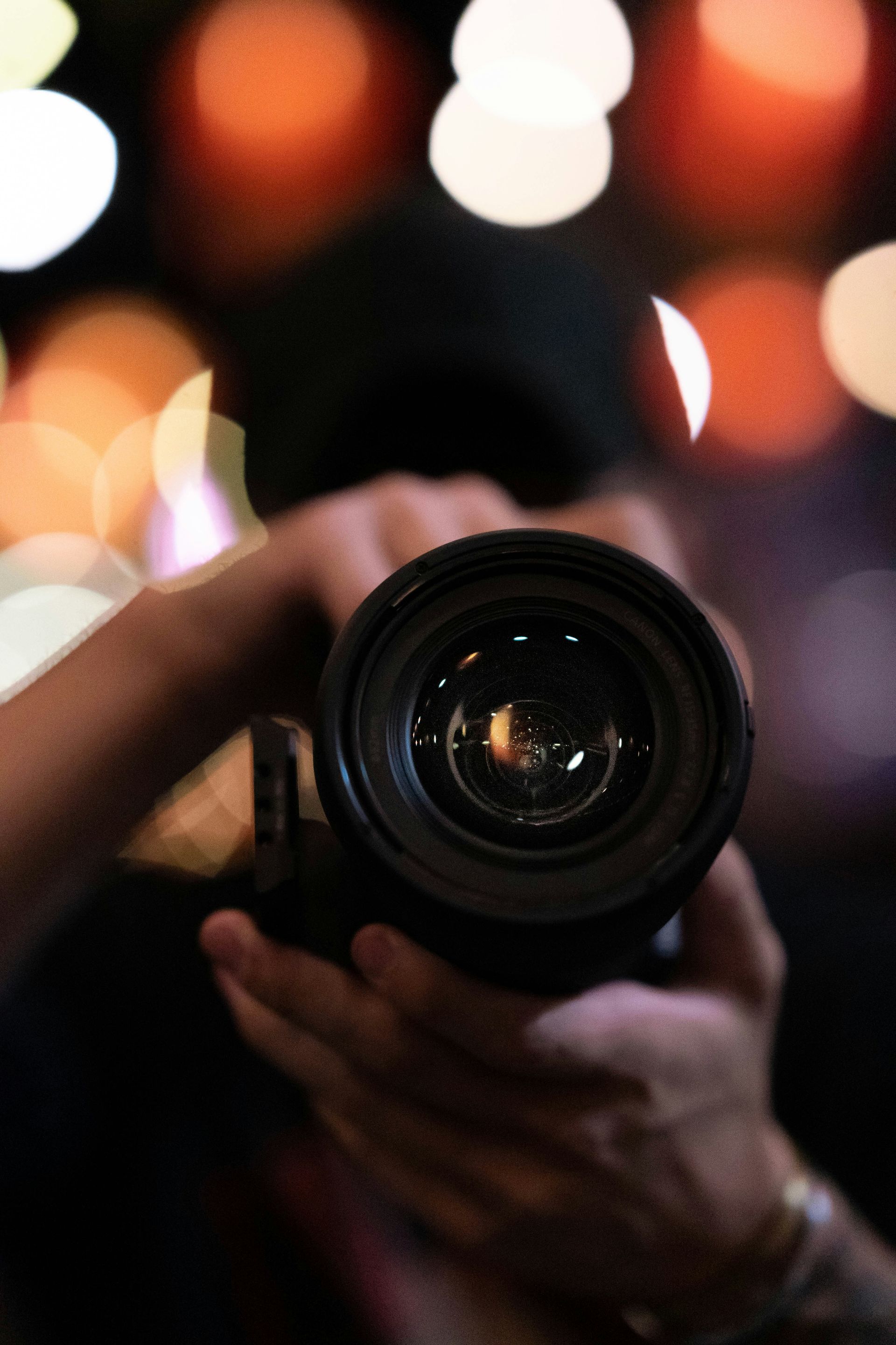 Photographer holding a camera, lens pointed towards the viewer, blurred background with orange and white bokeh.