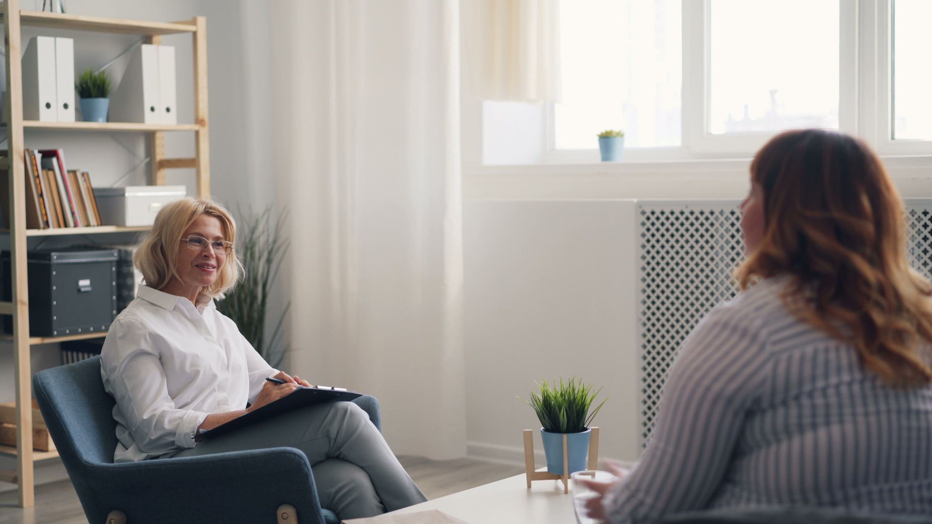 Therapist and client talking in a bright counseling office with bookshelves and a window.