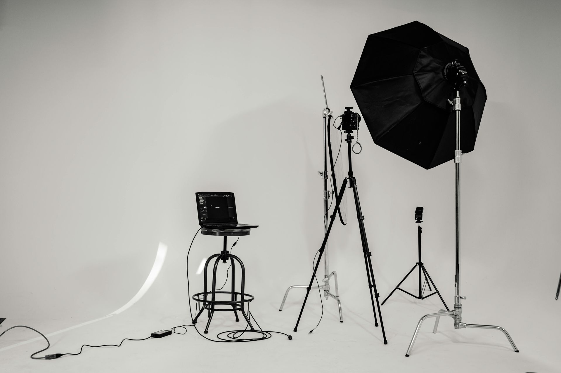 A photography studio setup with a laptop, lighting equipment, and a stool against a white backdrop.