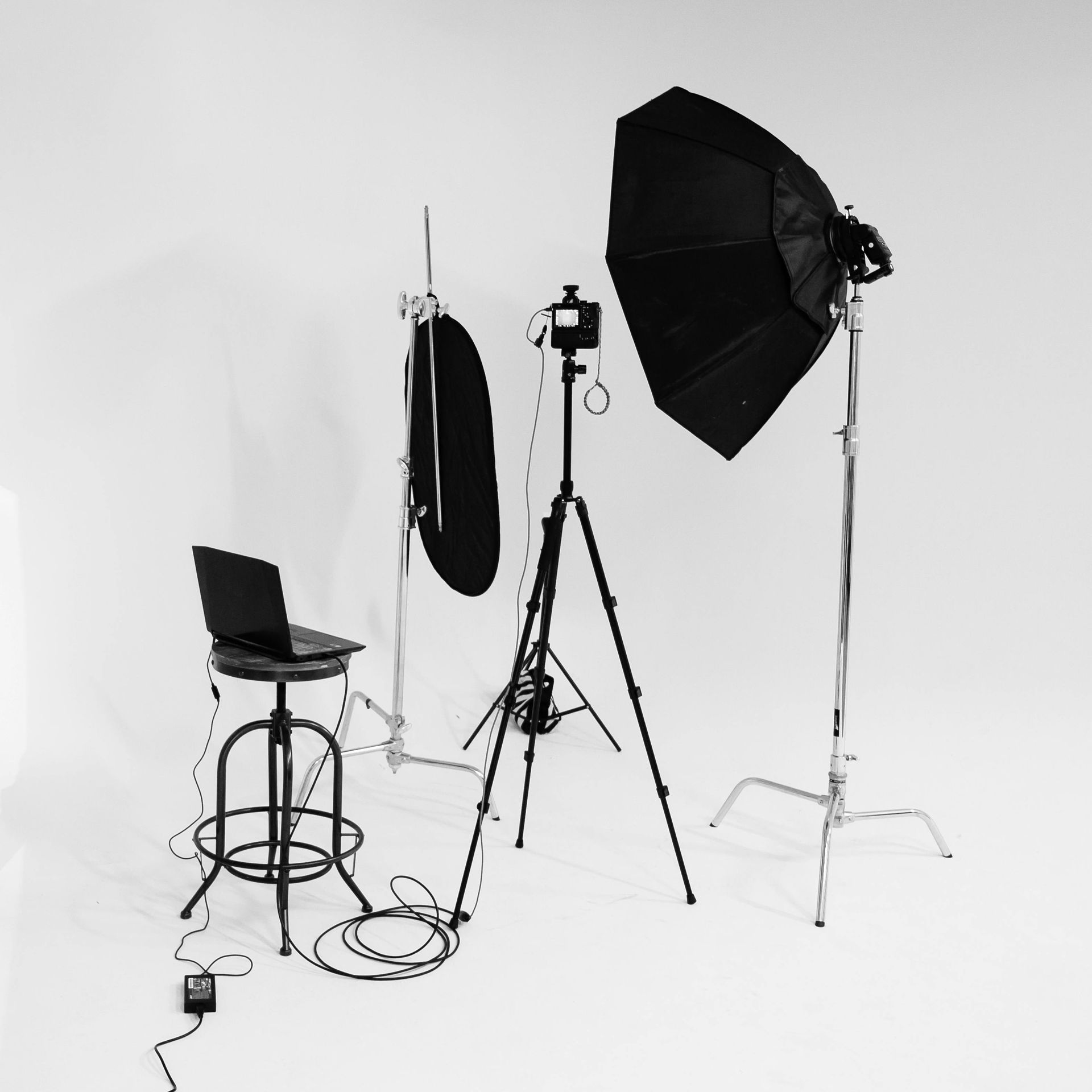 Black and white photo studio setup: stool with laptop, reflector, umbrella softbox, lights on stands.