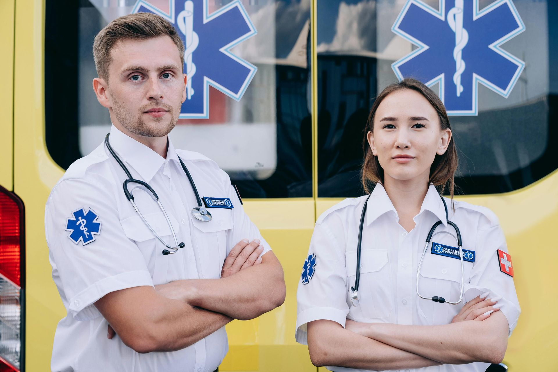 Two emergency medical workers in white uniforms stand in front of an ambulance, arms crossed.