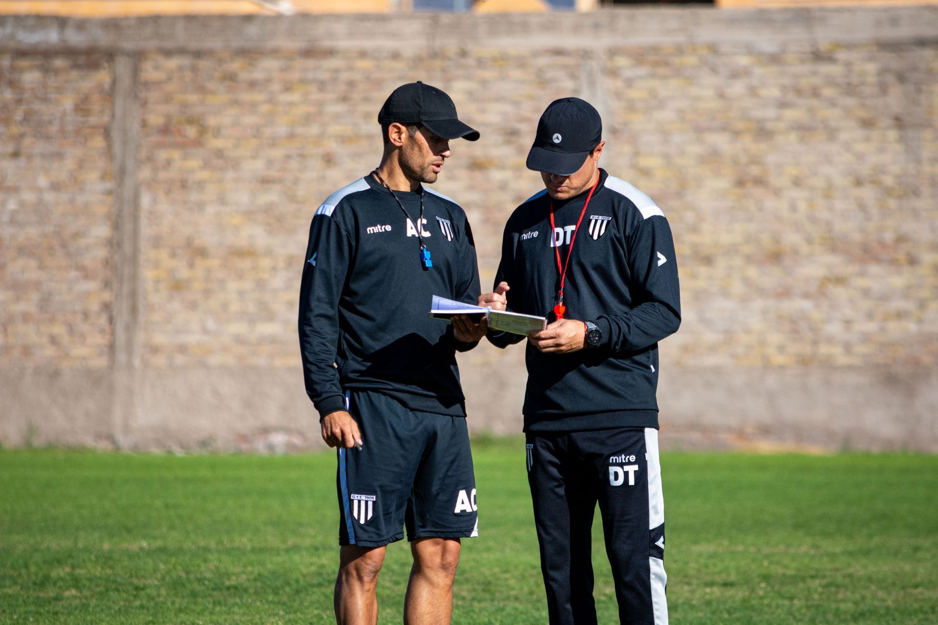 Two athletes in black training gear reviewing notes on a soccer field.