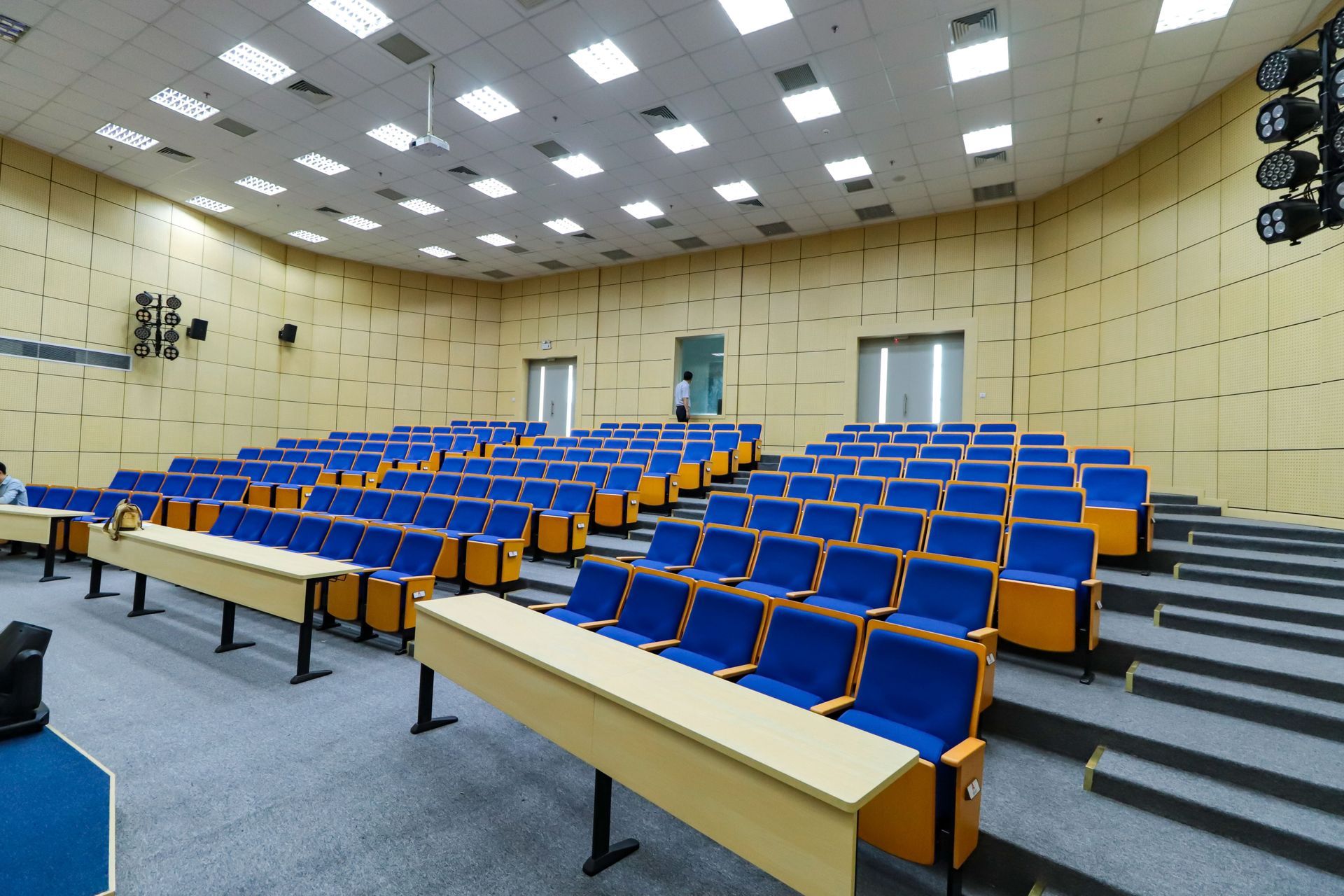 Empty lecture hall with blue seats, wooden desks, and a stage under bright ceiling lights