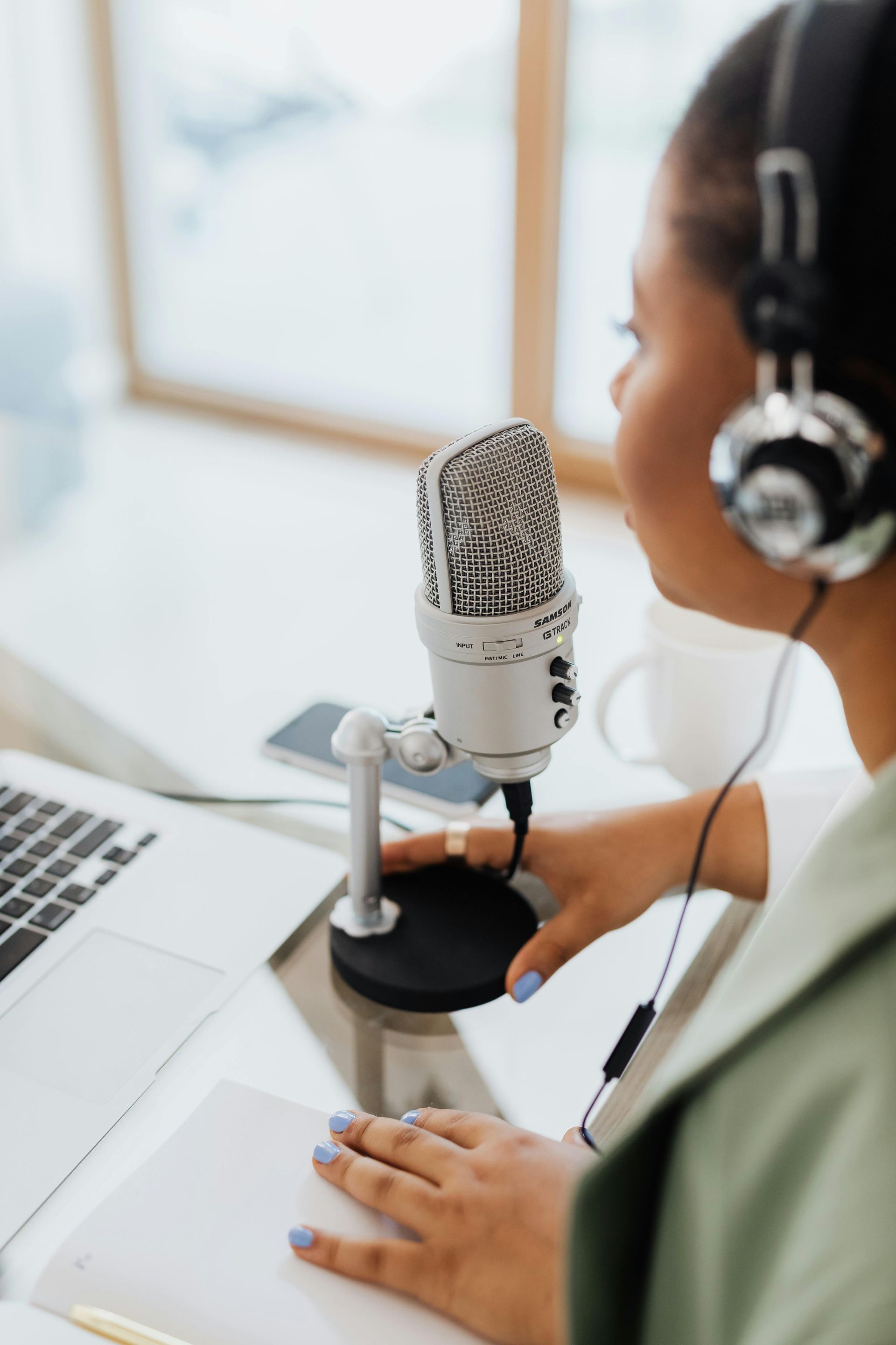 Woman with headphones recording podcast, seated at desk with microphone and laptop.