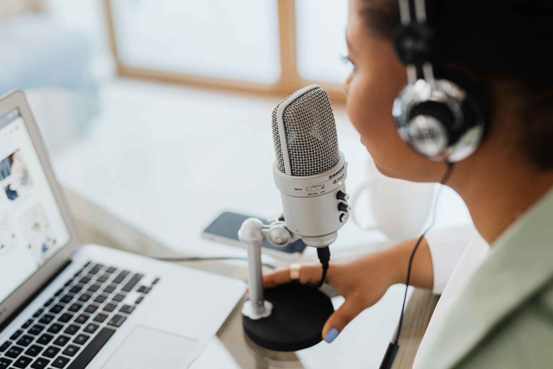 Woman with headphones, speaking into a microphone in front of a laptop, recording a podcast.