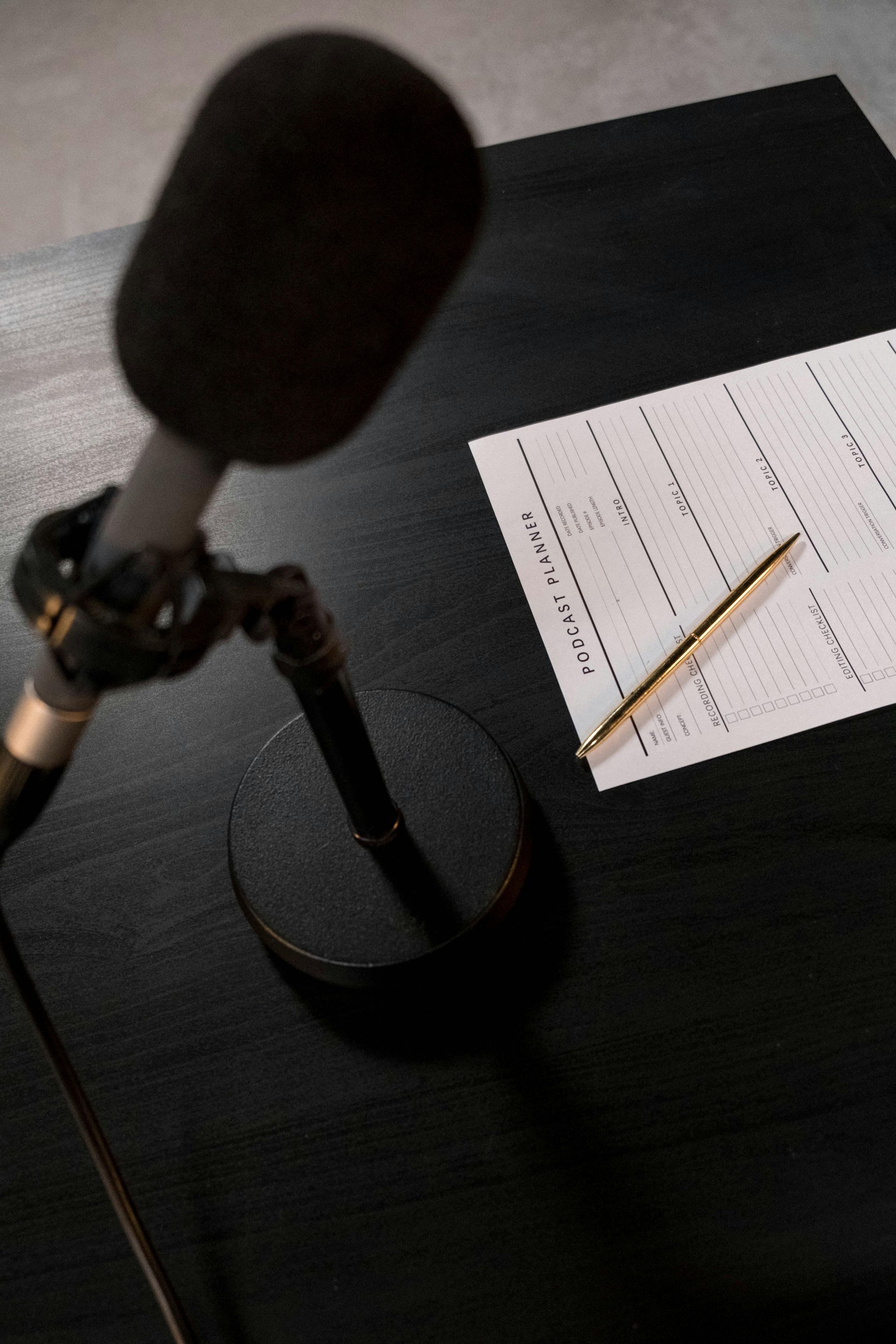 Microphone and script on a black desk, illuminated by shadows. A pen rests beside the paper.