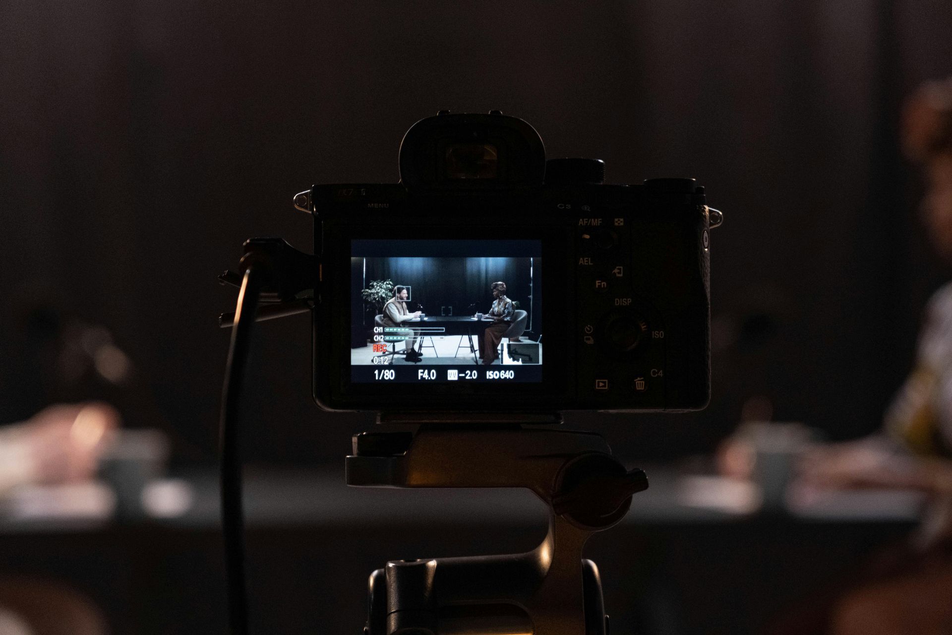 Camera on tripod records two people at a table in a dark studio; mics and cups present.
