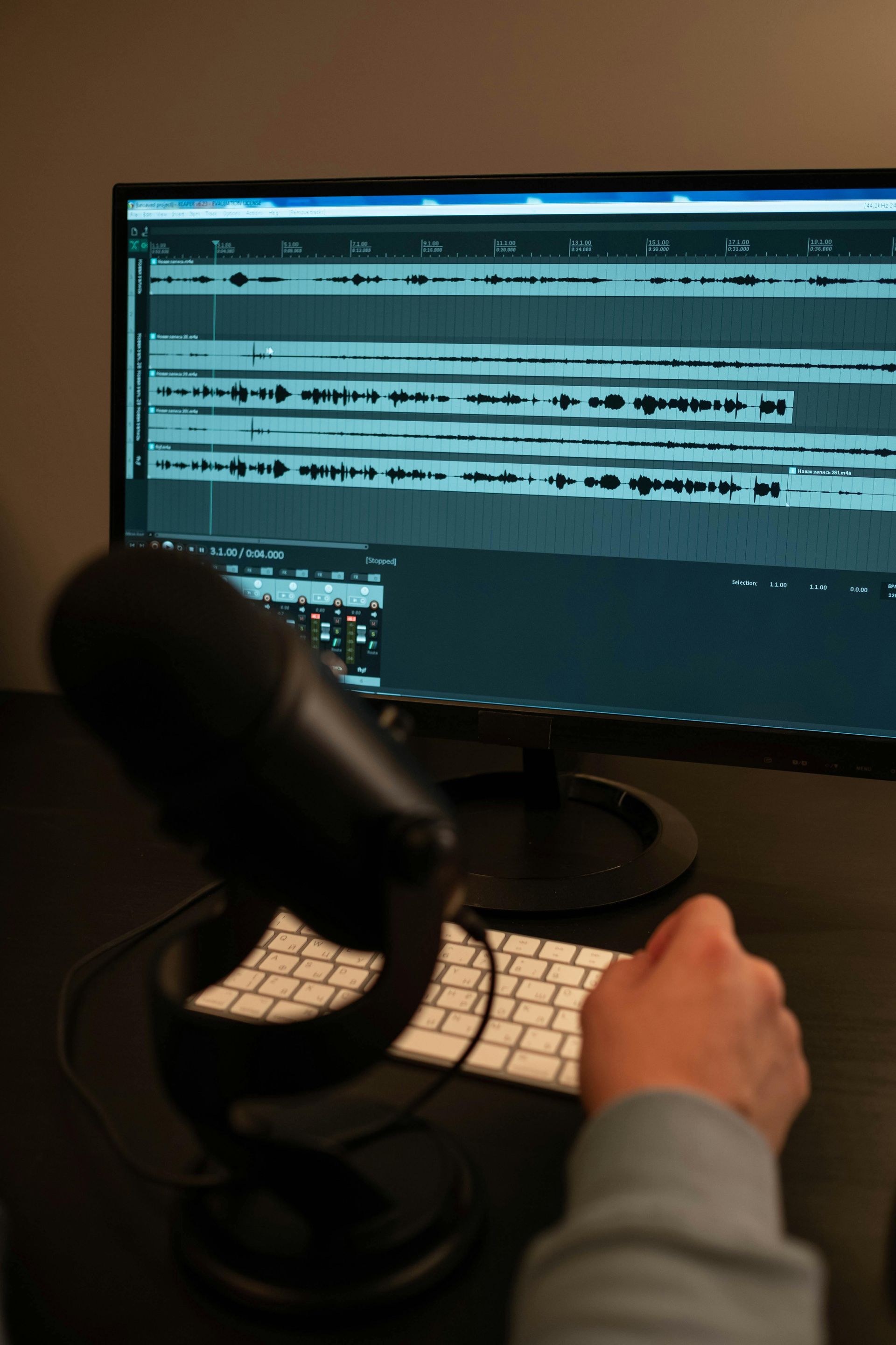 Microphone in front of computer monitor with audio editing software on screen. A hand is near the keyboard.