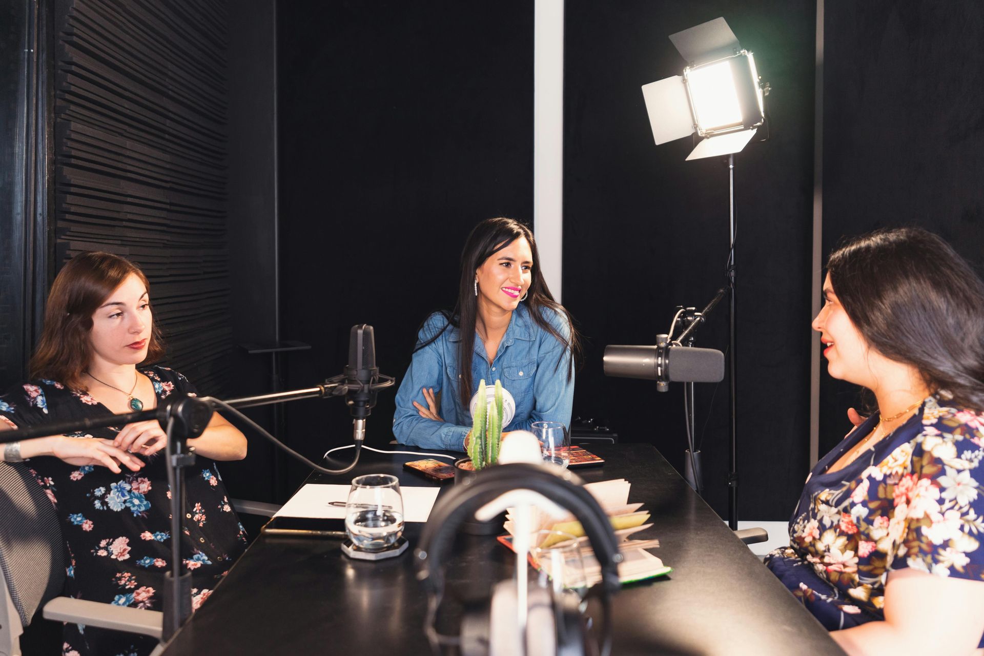 Three women seated at a table with microphones, podcast recording in a dark studio setting.