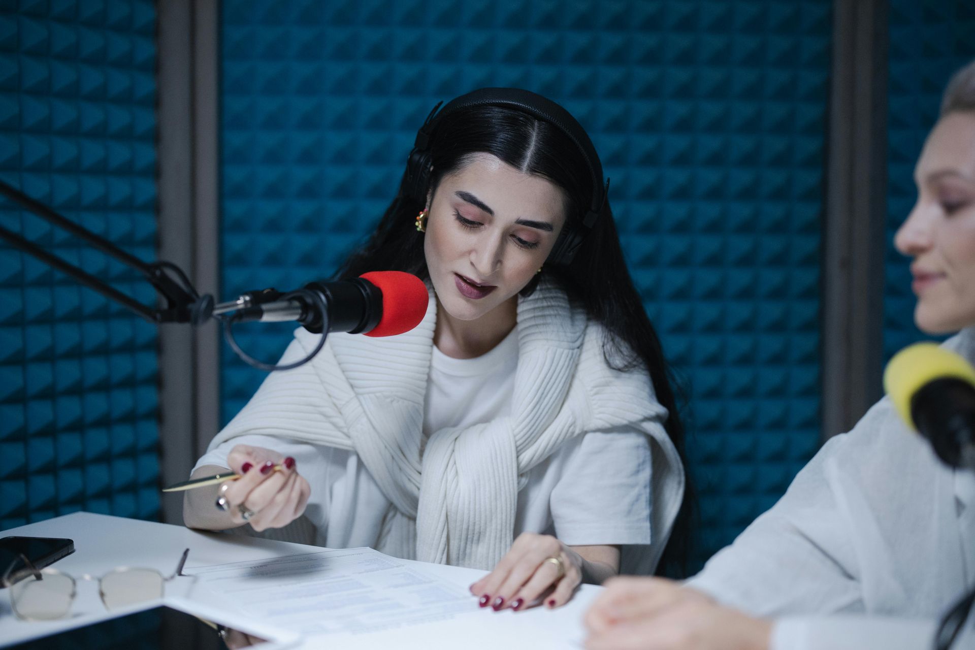 Two women at a desk in a recording studio. One speaks into a mic, pointing at notes.