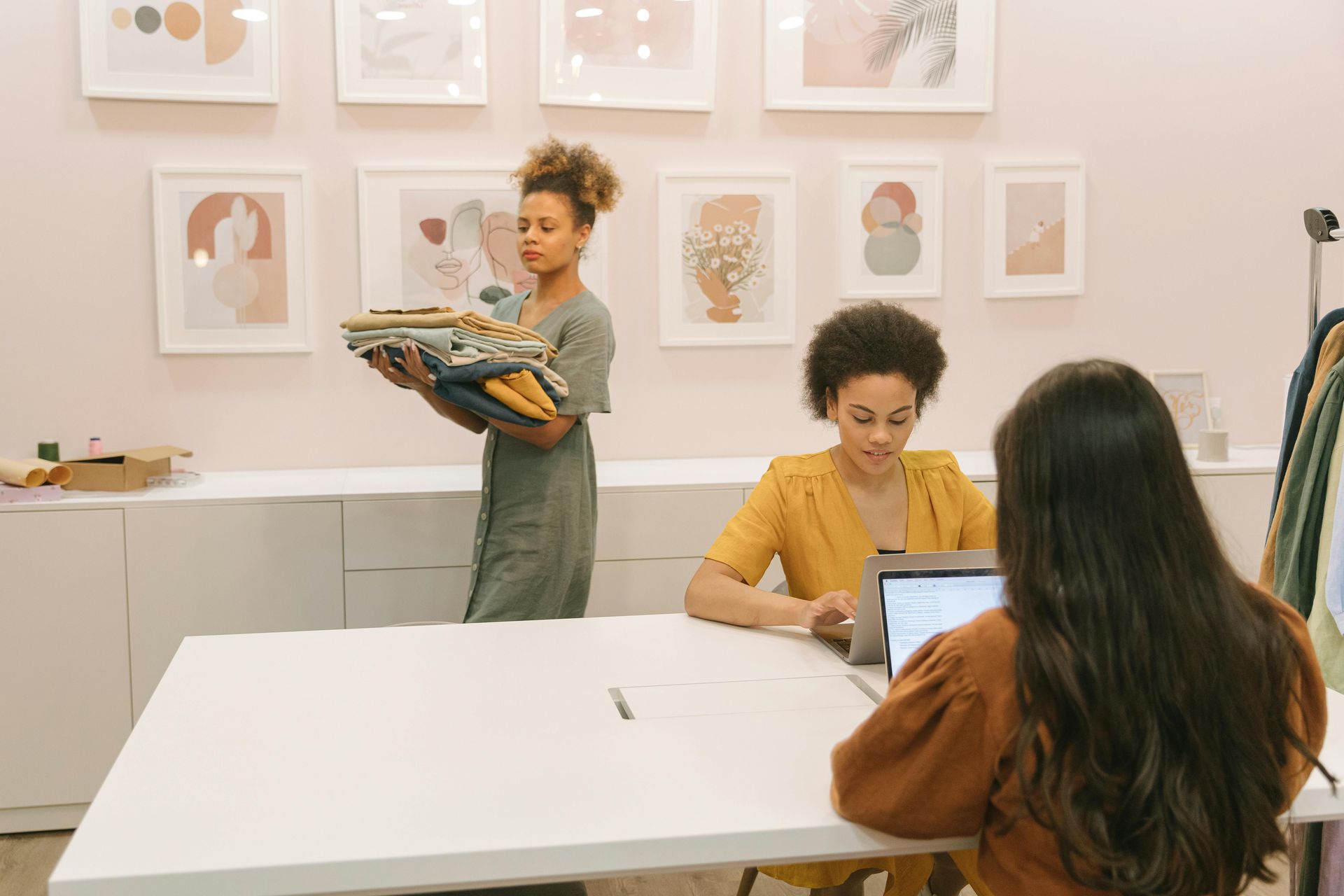 Two people in a bright art studio, one showing a sketchbook while another sits at a table with a laptop.