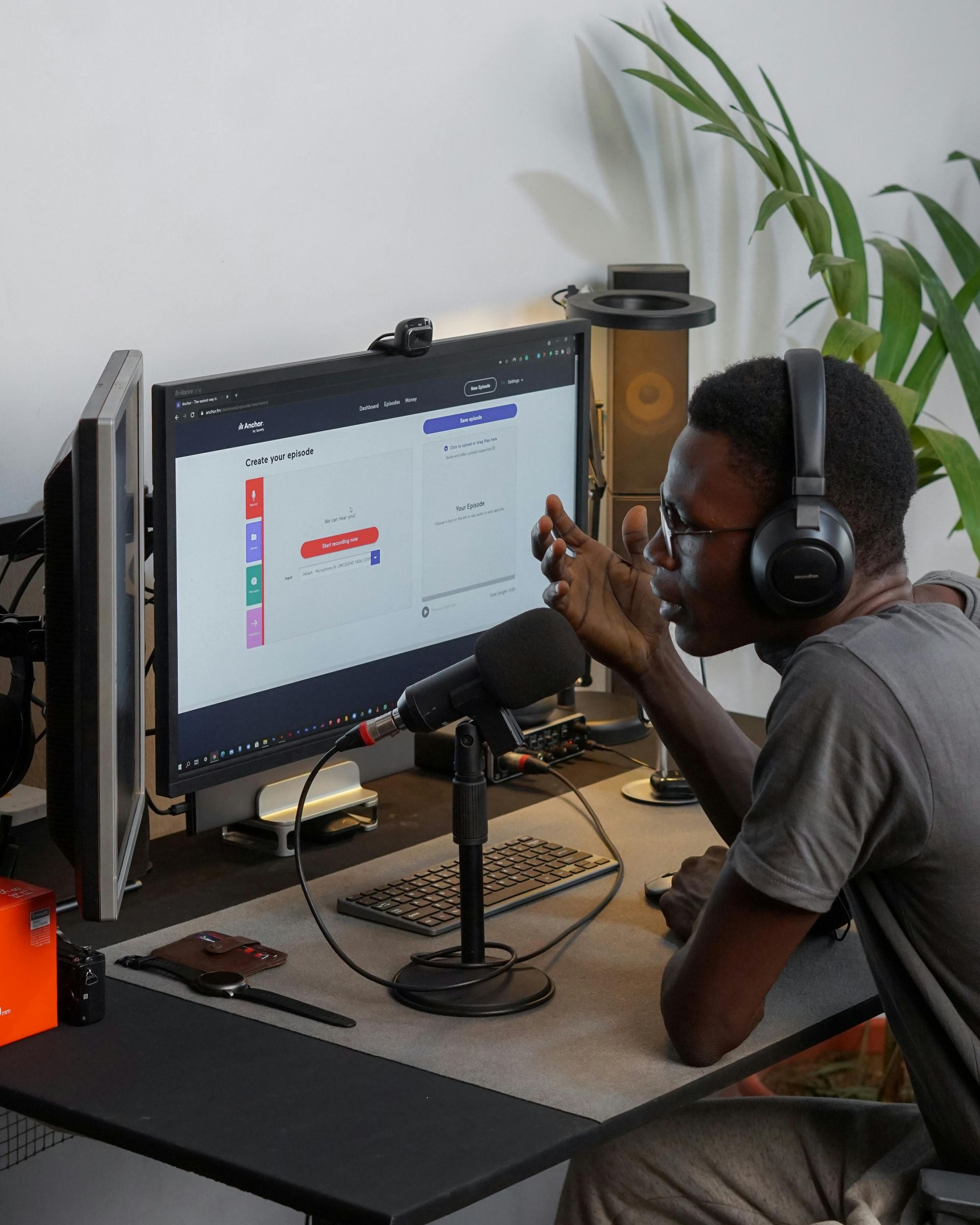 Man at computer with microphone and headphones, possibly podcasting. Dark desk, screen displaying graphs.