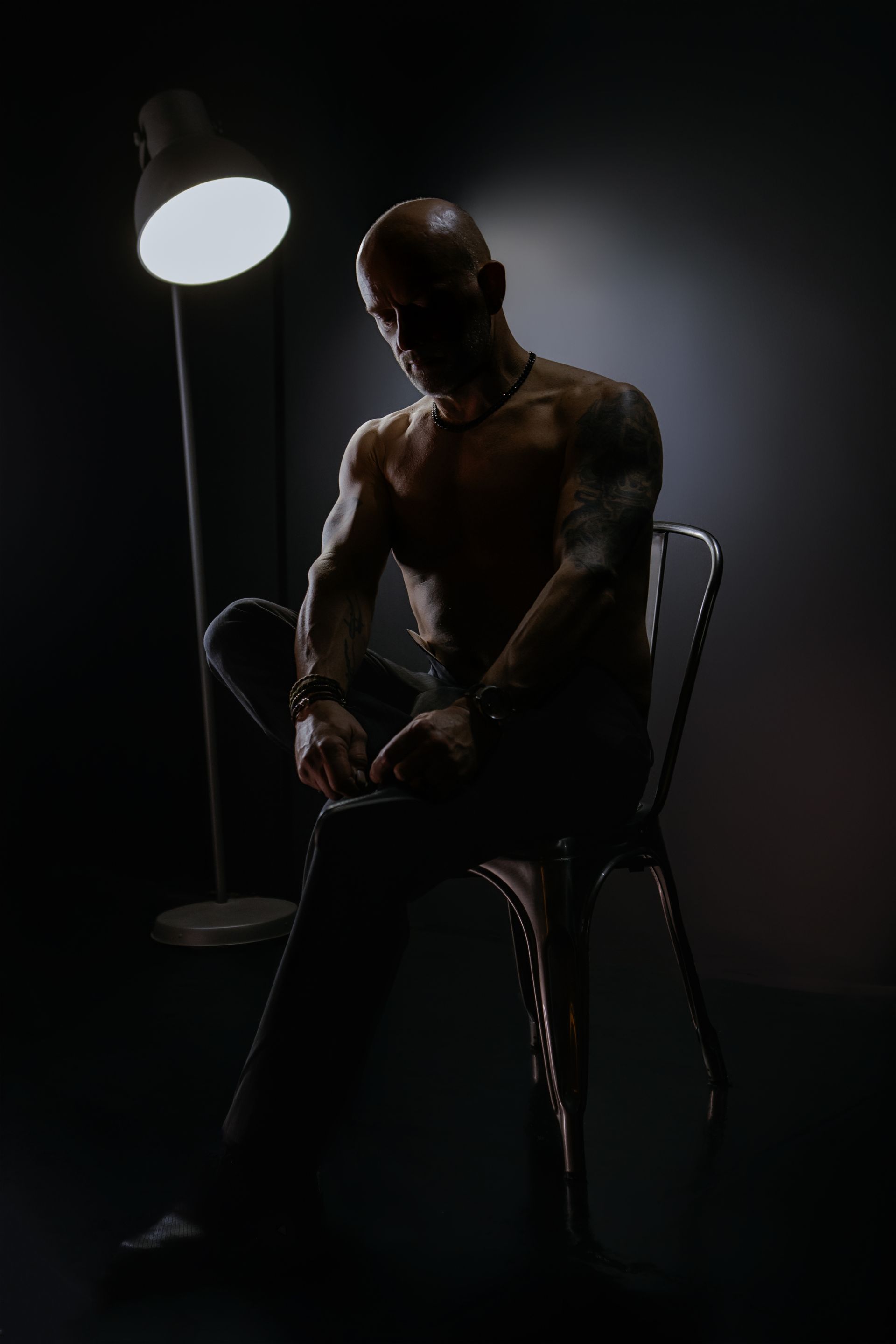 Dimly lit seated man in a dark room beside a glowing floor lamp, with visible tattoos.
