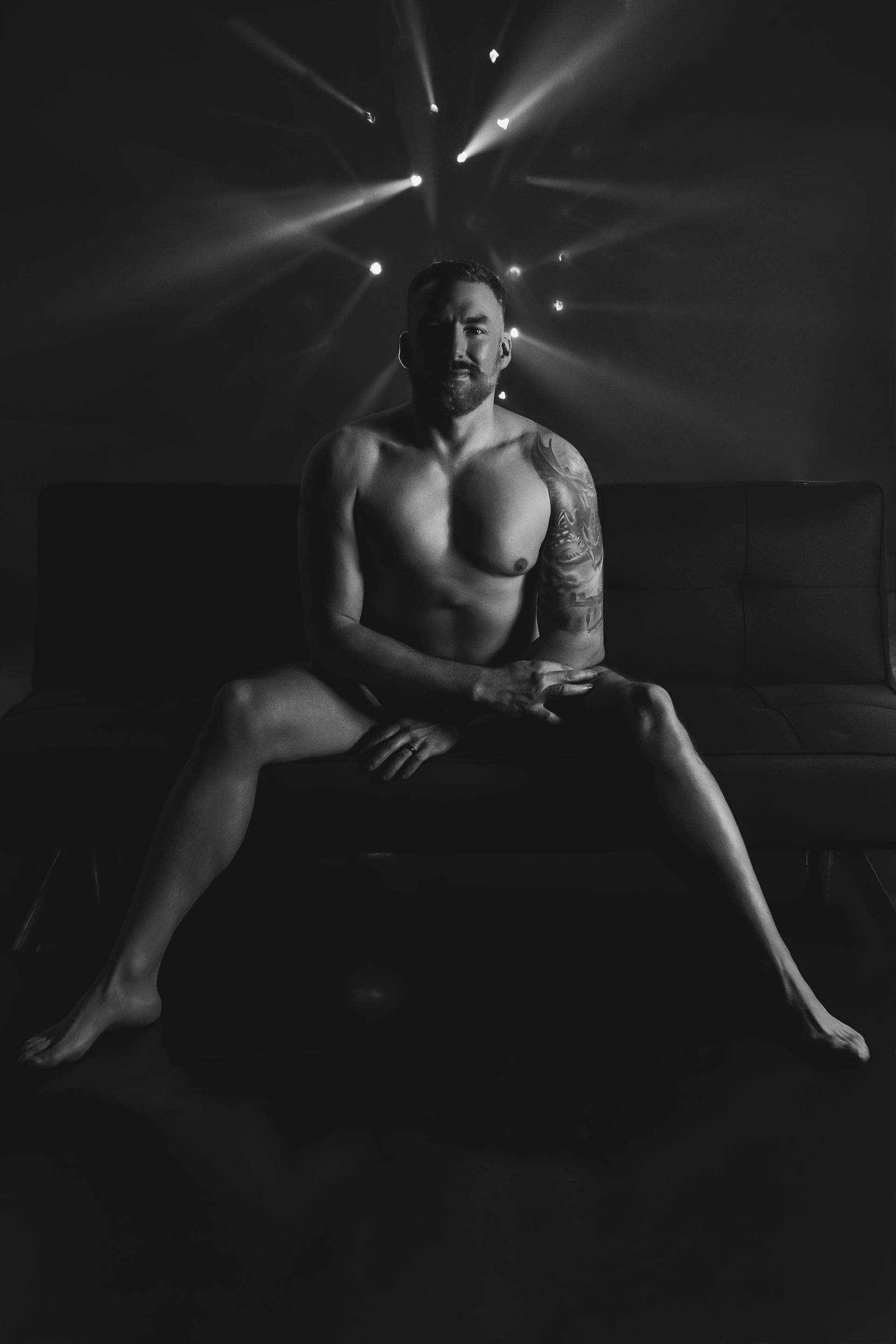 Black-and-white portrait of a shirtless person seated on a couch under dramatic backlighting