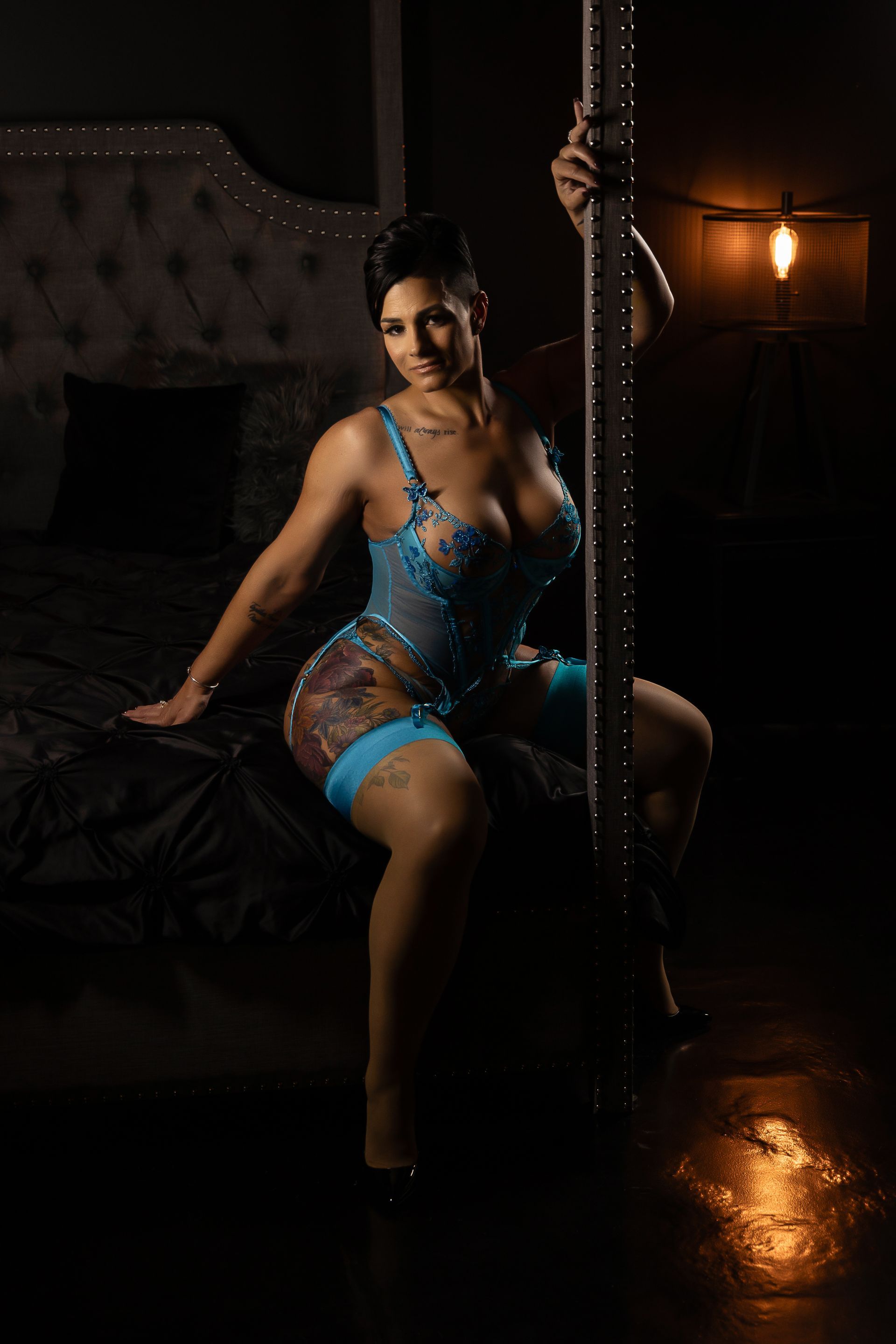 Woman seated on a pole in a dimly lit room, wearing a blue outfit with warm amber lighting.