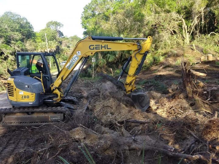 Yellow GEHL excavator digging dirt and debris in a wooded area, operator inside.
