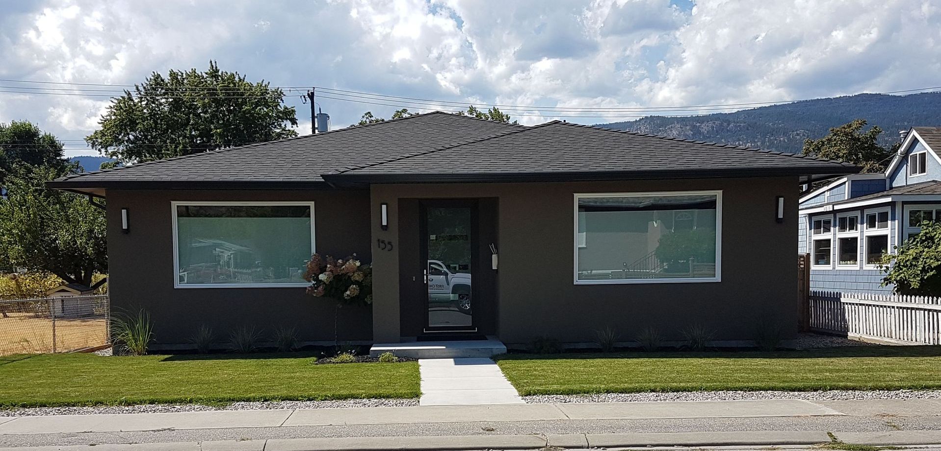 A modern, single-story, grey house with a white walkway and small front yard, in front of a road.