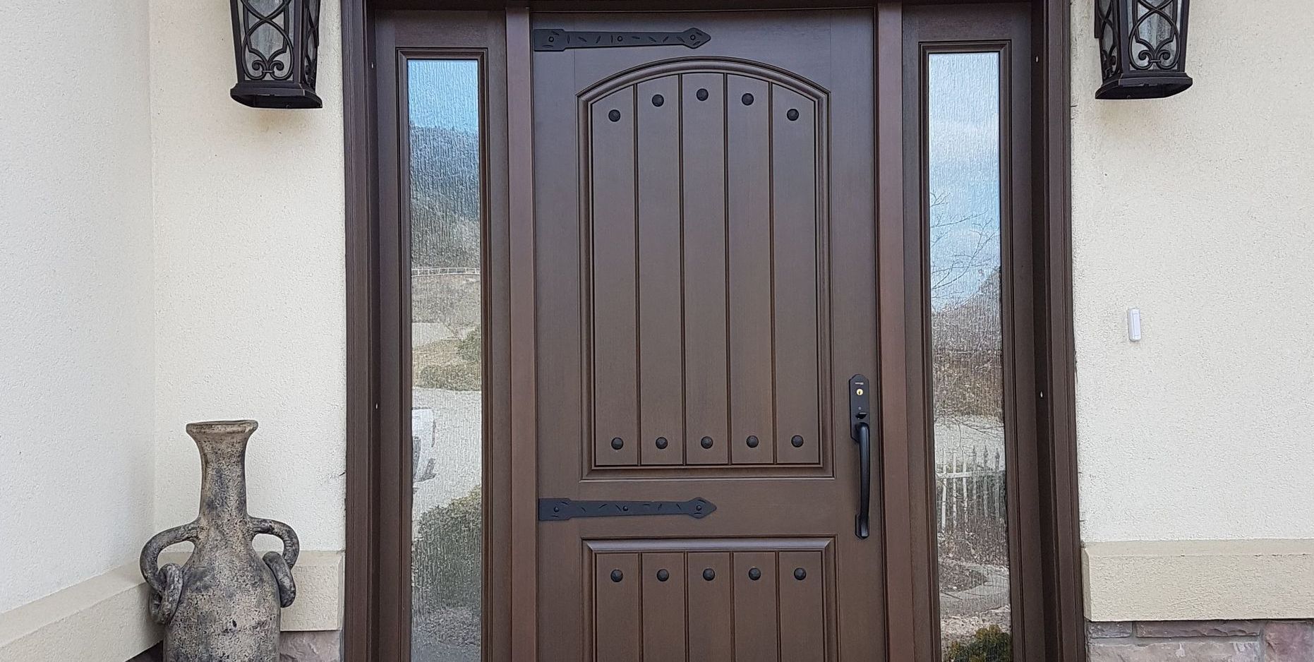 Brown wooden door with sidelights, decorative lanterns, and a vase on a stucco exterior.