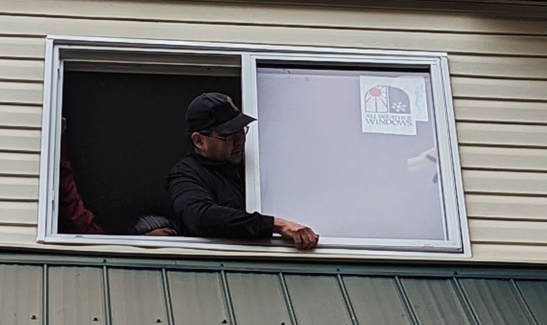 Man looks out a partially opened window. Gray siding and green trim of a building are visible.