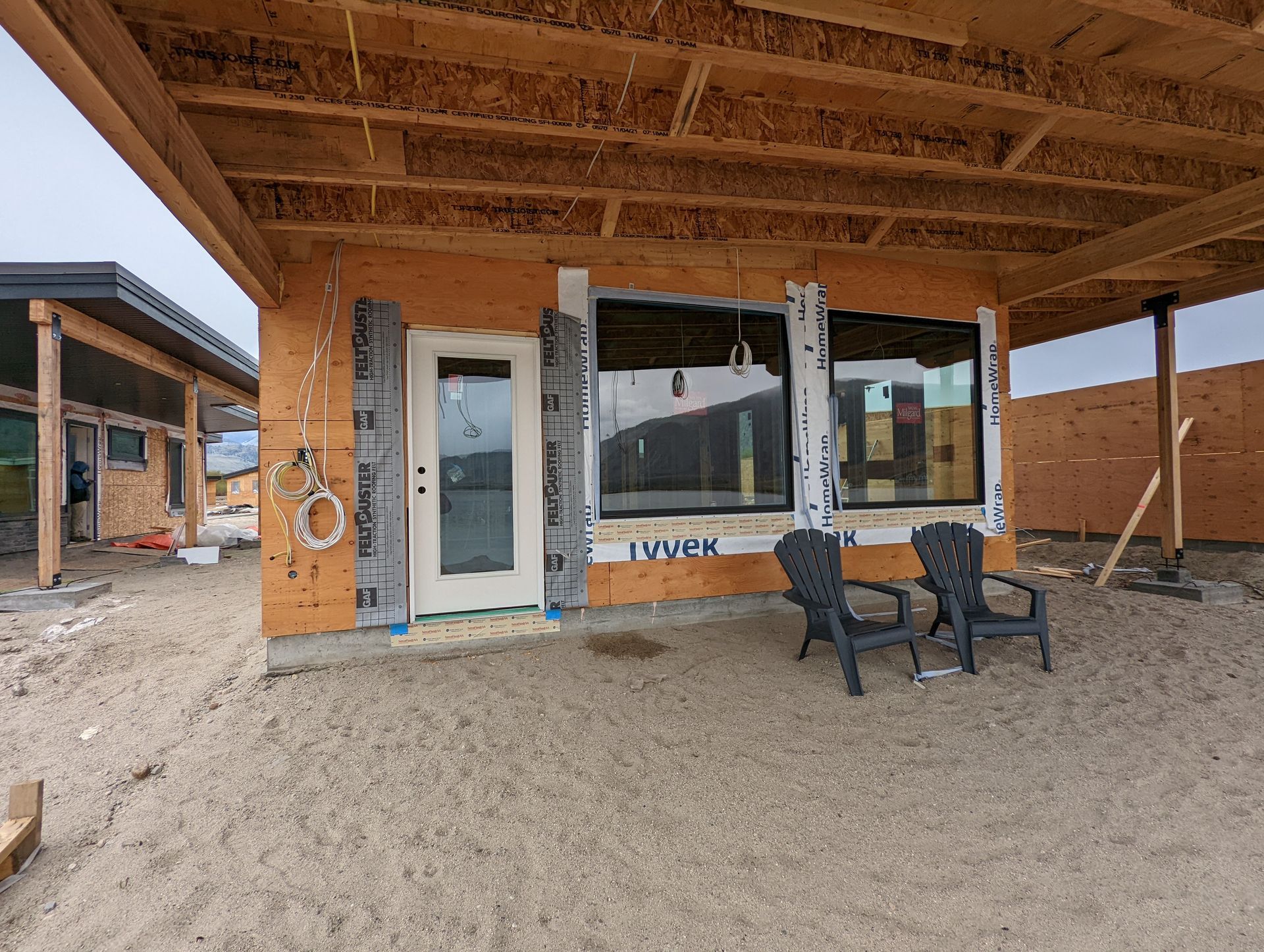 Construction site: a building with large windows and a door, two chairs on a sand area under a wooden overhang.