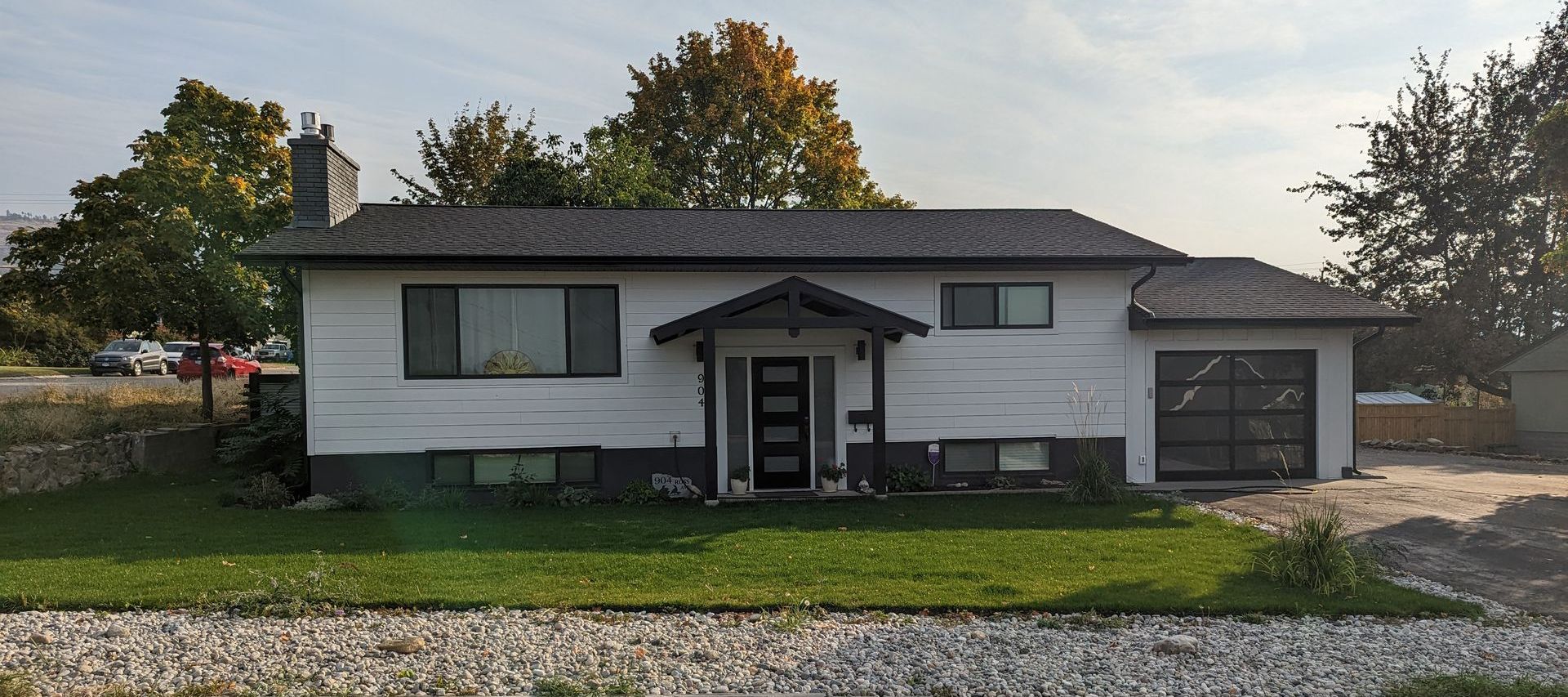 White house with black roof, windows, and garage door, trees on the right and left, green lawn, blue sky.