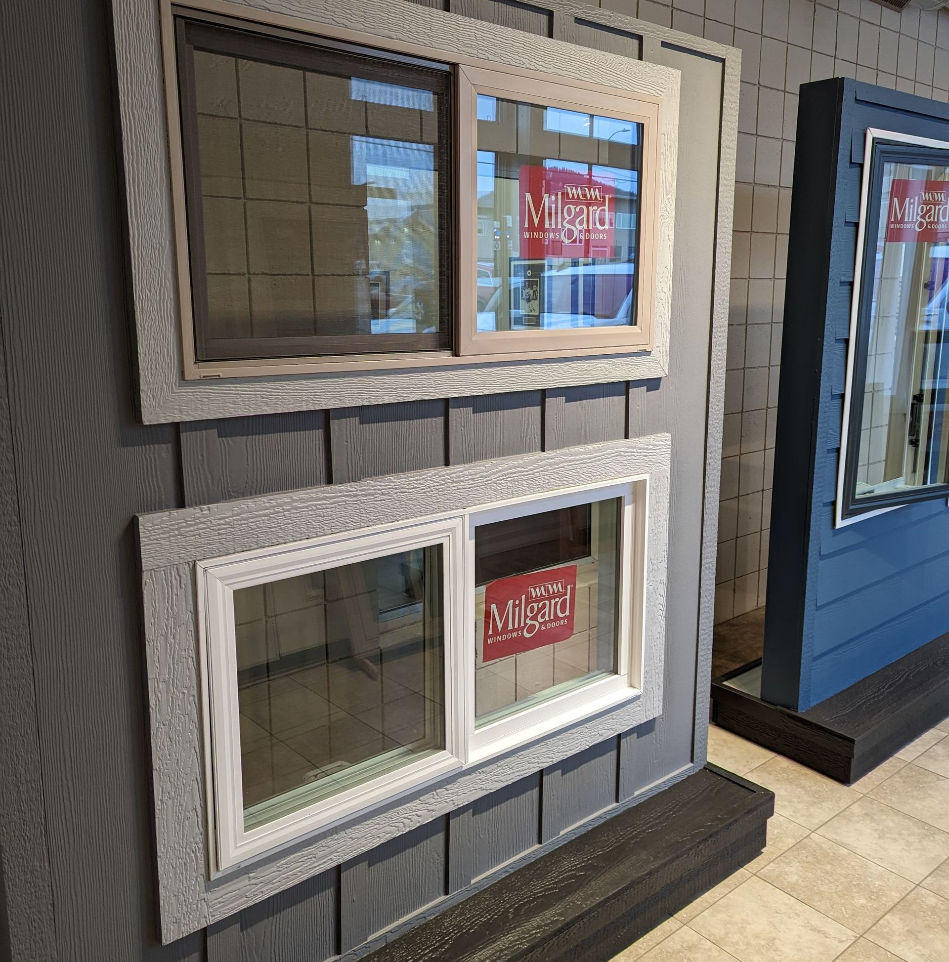 Two Millgard windows on display, set in a gray siding wall. One window is open, the other is closed.