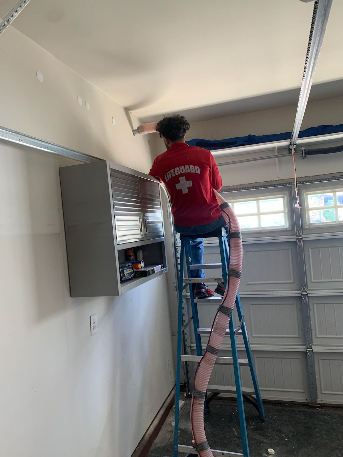 A man is sitting on a ladder in a garage painting the ceiling.