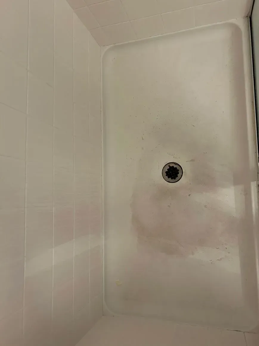 Top-down view of a white rectangular shower floor with dark discoloration and debris around the center drain.