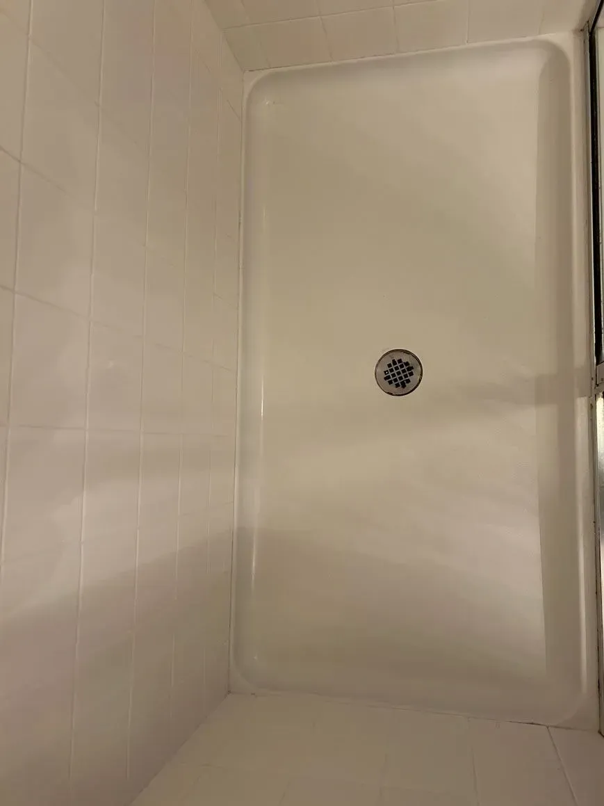 A white shower stall floor with a circular metal drain in the center, next to white tiled shower walls.