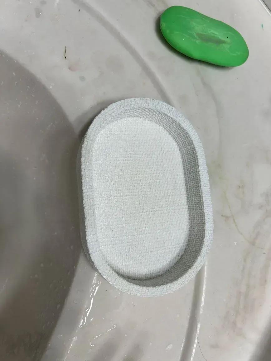 A white oval soap dish sits in a sink next to a small, green bar of soap.