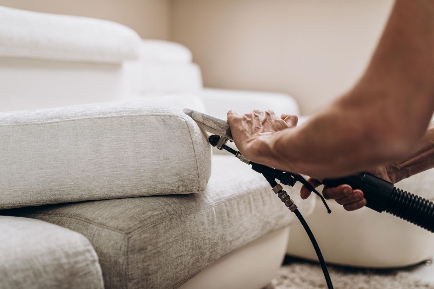A person uses a handheld upholstery cleaning tool to deep clean a textured, light-colored sofa.